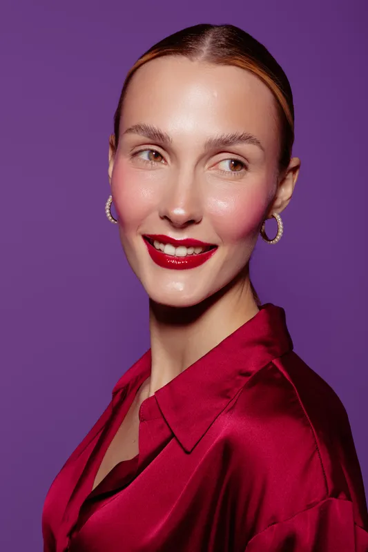Smiling woman with glossy red lips and hoop earrings looks sideways on purple background. She wears a silky red blouse. Concept of beauty, fashion, makeup, and elegance in studio.