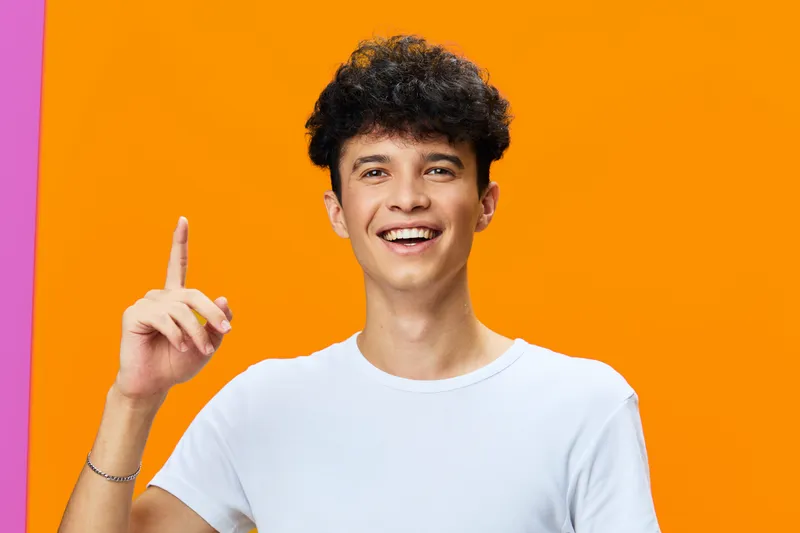 smiling man wearing white t-shirt pointing up with finger, cheerful young adult with curly hair and bright orange background, positive emotion and casual style