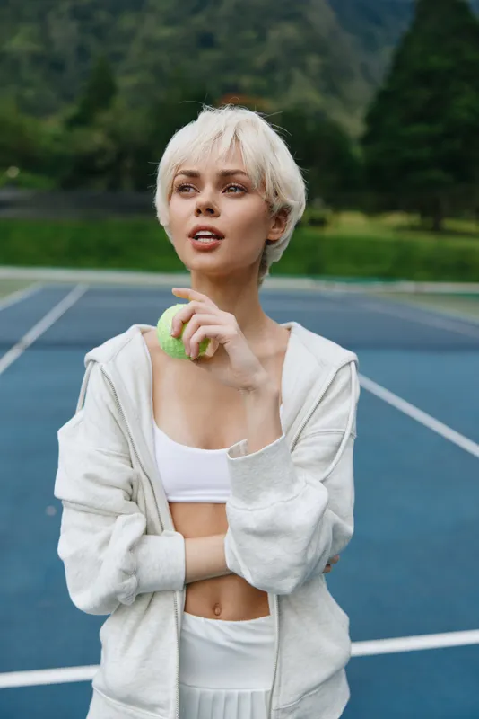 Young athletic woman with short blonde hair, dressed in a sports outfit, holding a tennis ball, enjoying a moment on a sunny outdoor tennis court