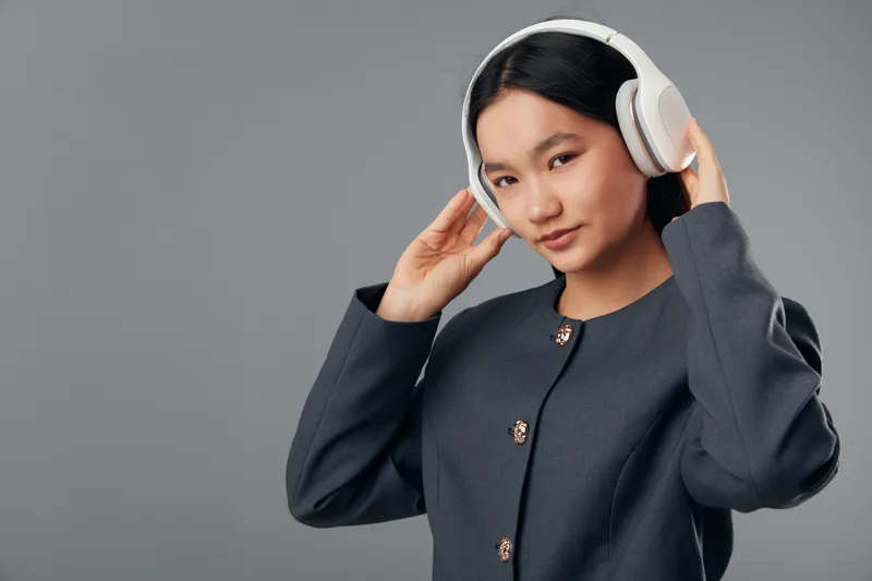 Woman with headphones, listening to music or audio content, smiling and posing on gray background. Young female with modern wireless headphones and casual dark jacket, entertainment concept.
