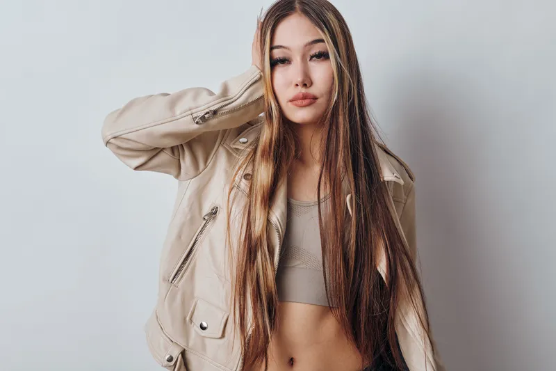 Young woman with long hair wearing beige jacket and crop top posing against white background. Fashion model with casual style and confident facial expression in studio.