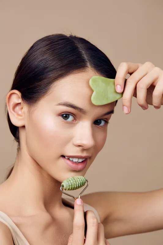 Young woman using jade roller and gua sha tool for facial massage, promoting skincare and wellness, against a soft beige background