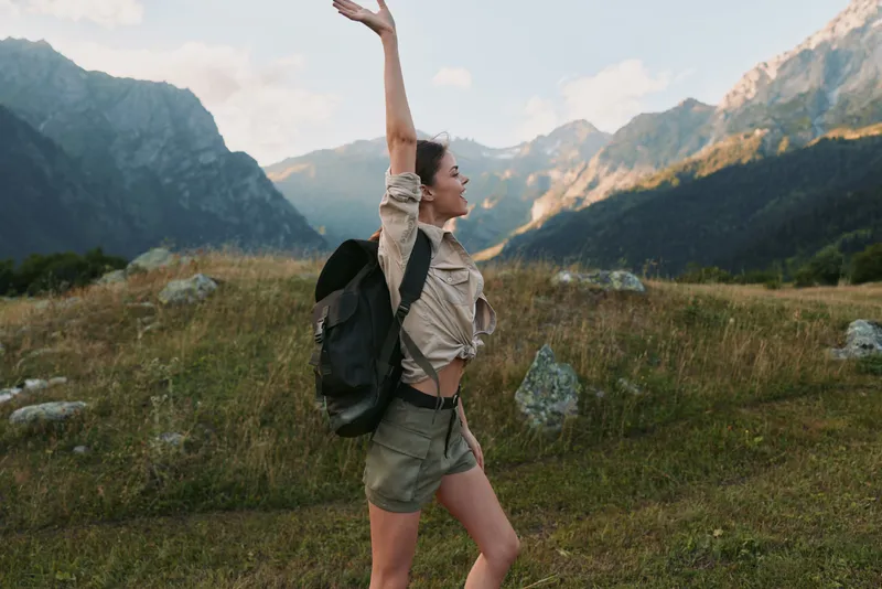 Happy woman hiking in mountains, raising arm in joy, wearing casual outfit with backpack, natural light, scenic landscape, outdoor adventure, lifestyle concept