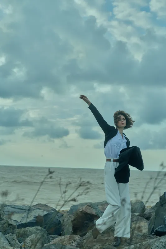 Outdoor scene of a female in white trousers and blazer on a rocky shore, wind in hair, reaching skyward for a dynamic coastal moment