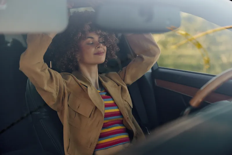 Young woman with curly hair sits behind the wheel in a sunlit vintage car, wearing a striped top and a tan jacket, enjoying a relaxed urban drive.