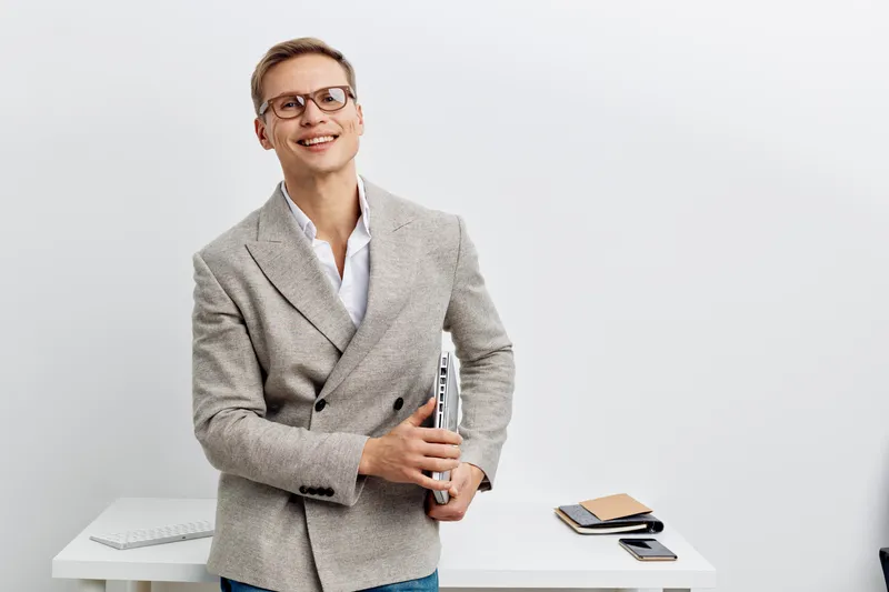 smiling man holding laptop with glasses wearing gray blazer standing office desk digital nomad concept