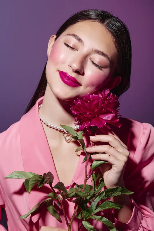 Young woman with closed eyes and bright pink lipstick holds dark pink peony flower close, wearing pink blazer and pearl necklace. Fashion and beauty creative portrait.