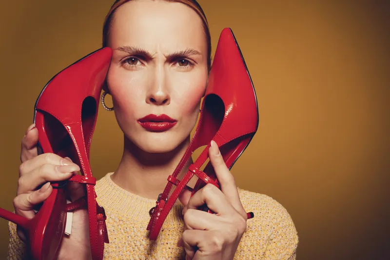 Woman with red lipstick holds red high heels near face with serious expression on yellow background. Concept for fashion, style, beauty, and footwear accessories.