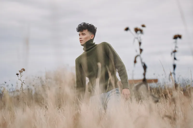 A young man in a green knit turtleneck walks through tall dry grass, captured in a candid, authentic portrait style conveying genuine presence, natural texture, subtle movement and introspective mood.