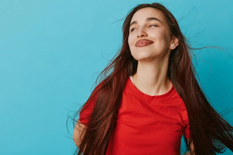 Young woman with long dark hair and red shirt smiling with closed eyes on blue background. Portrait of happy female enjoying moment with natural expression and carefree mood.