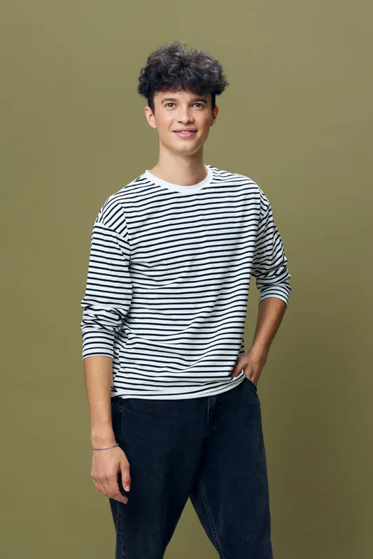 smiling man wearing striped long sleeve shirt stands against olive background, casual style, relaxed pose, black hair, young adult, natural lighting