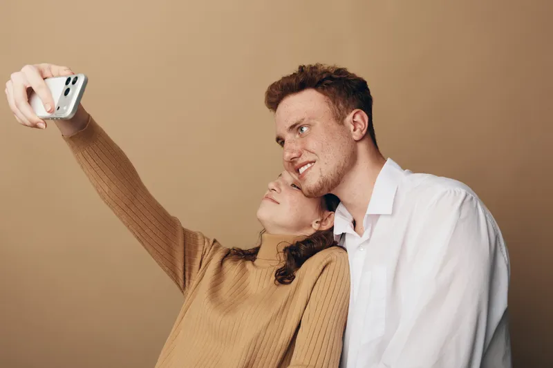 Young couple takes a selfie together on a beige background. The woman in a brown sweater leans on the man in a white shirt. Concept of friendship, love, and casual lifestyle.