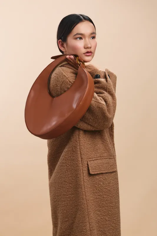 Asian woman wearing a textured coat poses with a circular brown bag over her shoulder in a studio setting, conveying a chic, urban fashion mood.