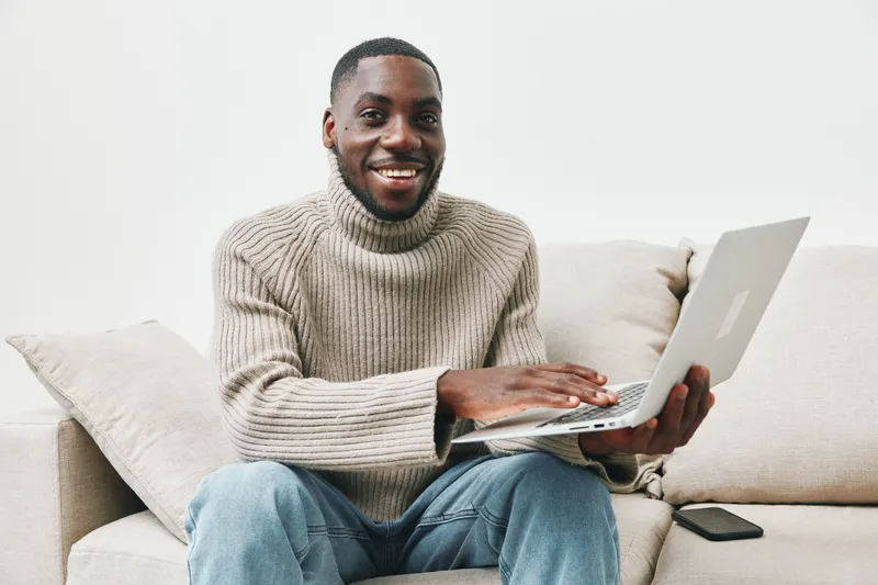 Young African man sitting on a sofa, smiling while working on a laptop, wearing a cozy turtleneck sweater, embodies comfort and productivity in a warm, minimalistic setting