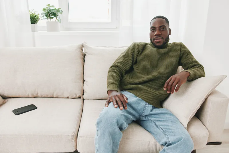 Young African man relaxing on a beige sofa in casual green sweater and blue jeans, looking calm and confident, in a bright modern living room