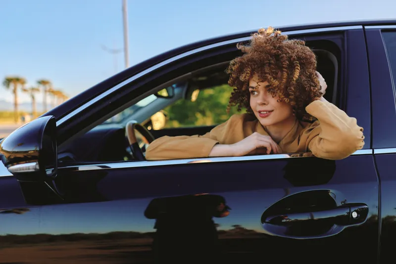 Curly haired woman leaning out of a sleek car window, casual lifestyle vibe, retro film color, warm sunlight, relaxed driving pose, celebrating freedom on open road