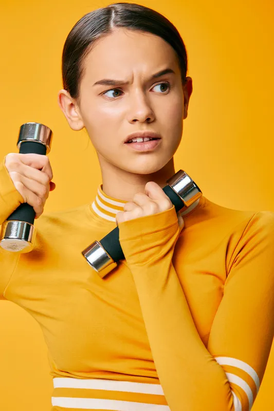 Woman fitness athlete in bright orange athletic top holding dumbbells, focused expression, yellow studio backdrop conveying strength, effort and athletic determination during a training session.
