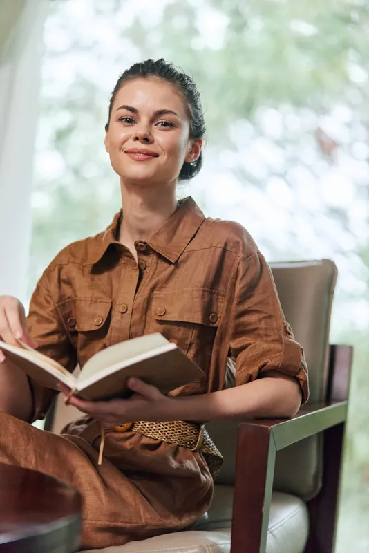 Молодая женщина в льняном платье коричневого цвета улыбается, читая книгу в уютной обстановке