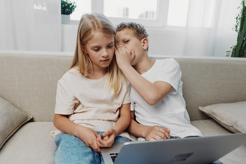Boy whispers secret to girl sitting on sofa with laptop, casual kids sharing private moment at home with natural light and neutral interior background.