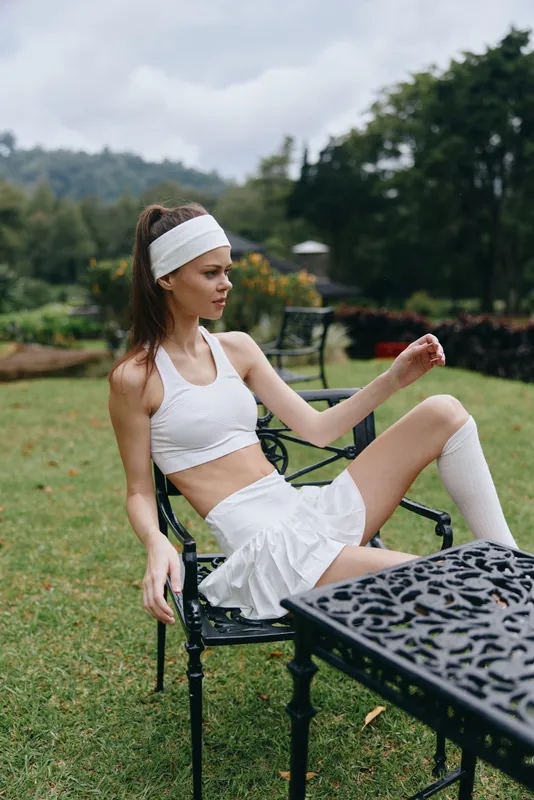 Fitness model in white activewear relaxing on a chair outdoors, showcasing a sporty lifestyle amidst lush greenery and cloudy skies