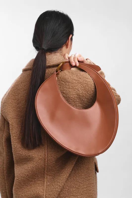 asian woman wearing a warm coat, back turned with a round leather bag over the shoulder, seen in a clean studio setting, projecting stylish confidence and poised elegance.