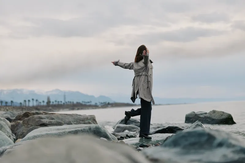 Woman in casual clothing stands on rocks by the sea with arms outstretched, facing cloudy sky. Concept of freedom, nature, and peaceful outdoor moment by the coast.