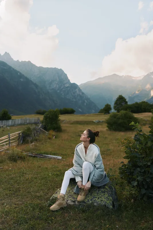 A young woman sits on a rock in an open meadow with distant mountains, looking up thoughtfully as soft light falls across the landscape.
