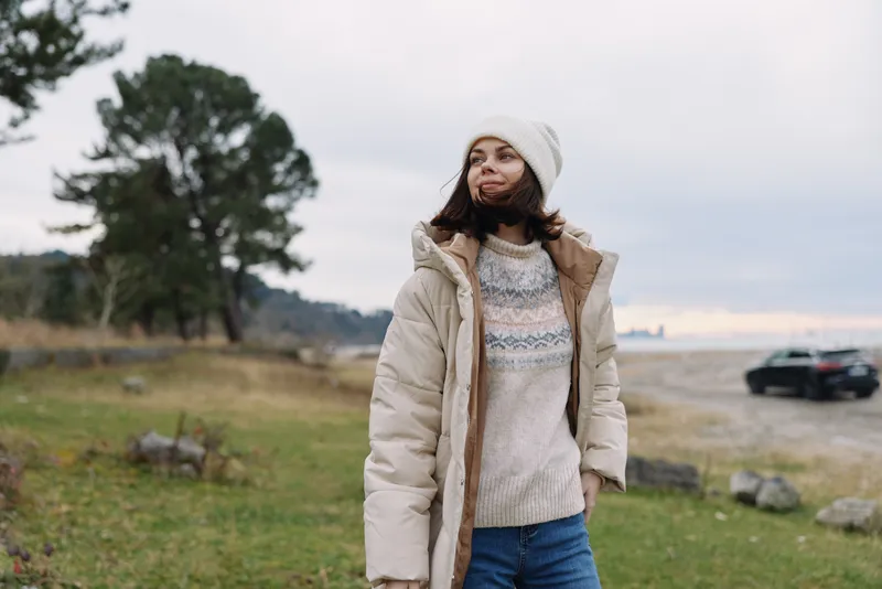 Woman outdoor winter clothing jacket sweater beanie standing in a windswept coastal field with trees and distant sea, casual lifestyle shot showcasing cold weather fashion