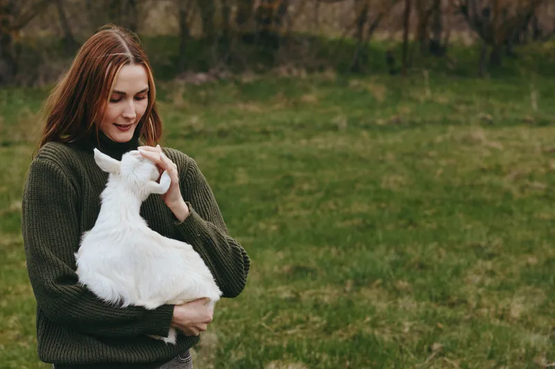 Casual woman outdoors cradles a white goat kid, smiling softly in a green field. The scene conveys a gentle lifestyle and close bond with animals, capturing calm rural moments.