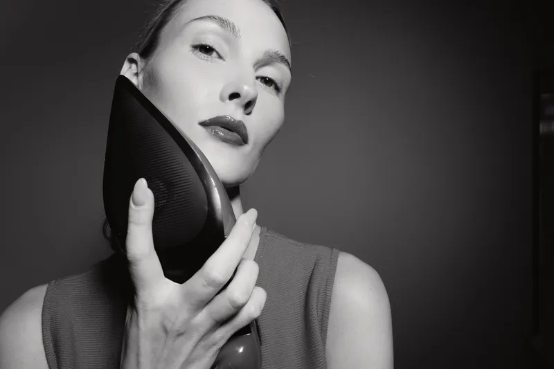 White woman with light skin holds black high heel shoe close to face in black and white studio portrait. Concept of fashion, elegance, and beauty.