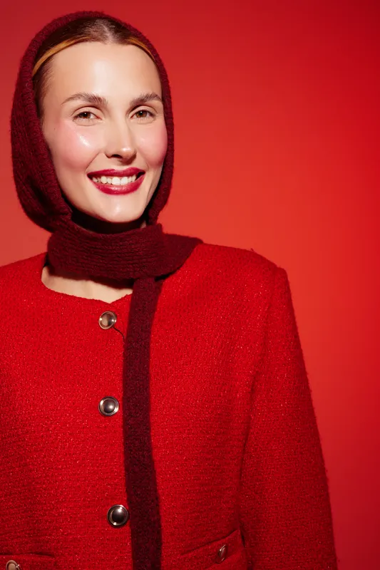 Smiling woman in red coat and matching scarf poses against red background. Bright makeup and glossy lips highlight her joyful expression and warm winter fashion style.