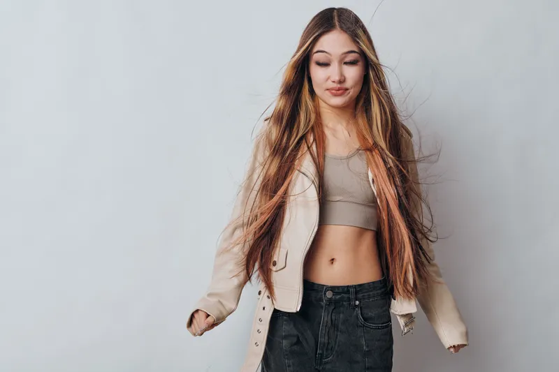 Young woman with long hair wearing beige cropped top and light jacket standing against gray background, casual style, confident pose and natural makeup for lifestyle portrait.
