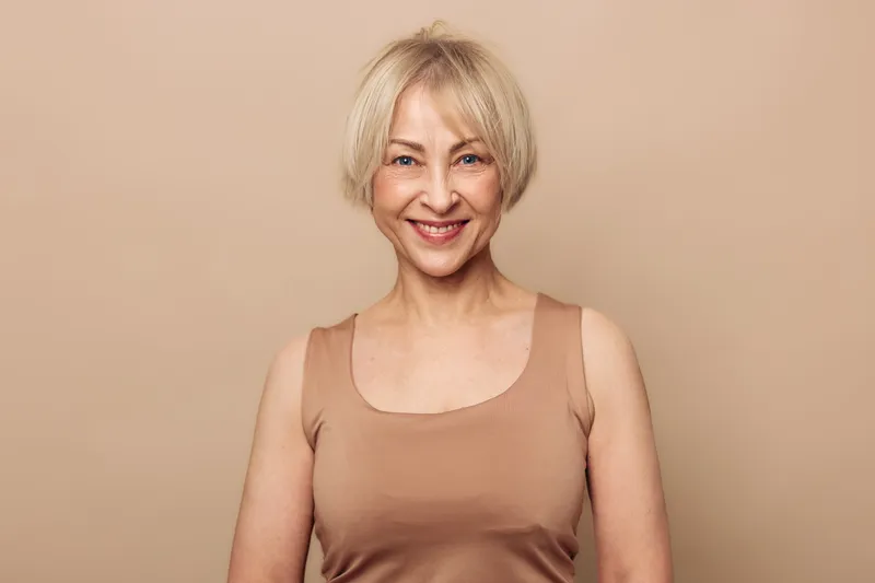 Smiling mature woman with short blonde hair wears beige tank top against light brown background. Concept of natural beauty, confidence, aging gracefully and positive lifestyle.