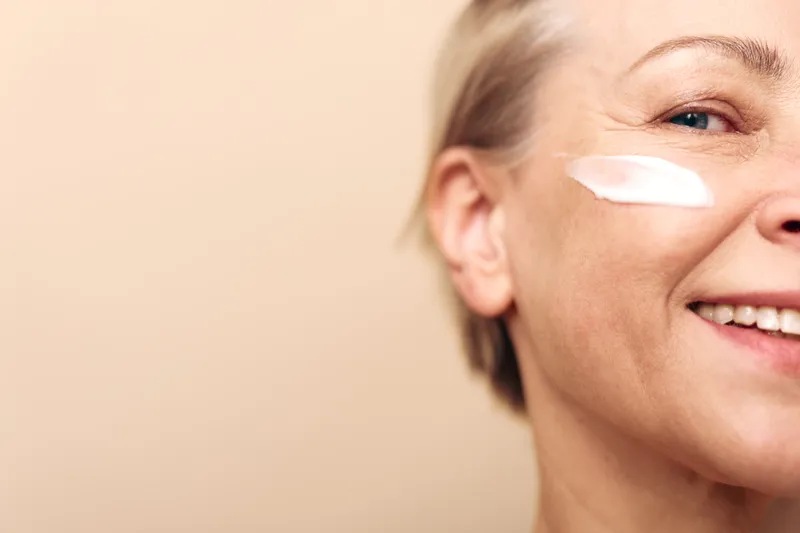 Close-up of smiling middle aged woman with cream on cheek against beige background. Concept of skincare, anti aging, self care, and natural beauty in wellness routine.