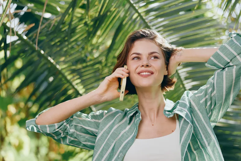 Portrait of a smiling woman talking on a cellphone outdoors, bathed in bright sunlight, framed by lush tropical greenery.