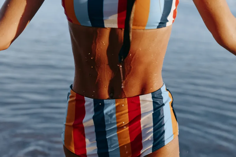 Woman bikini striped sea waves summer swimsuit vacation concept, close up of legs and torso on a beach with ocean ripples, warm sunlight highlights skin and pattern. Relaxed standing posture suggests