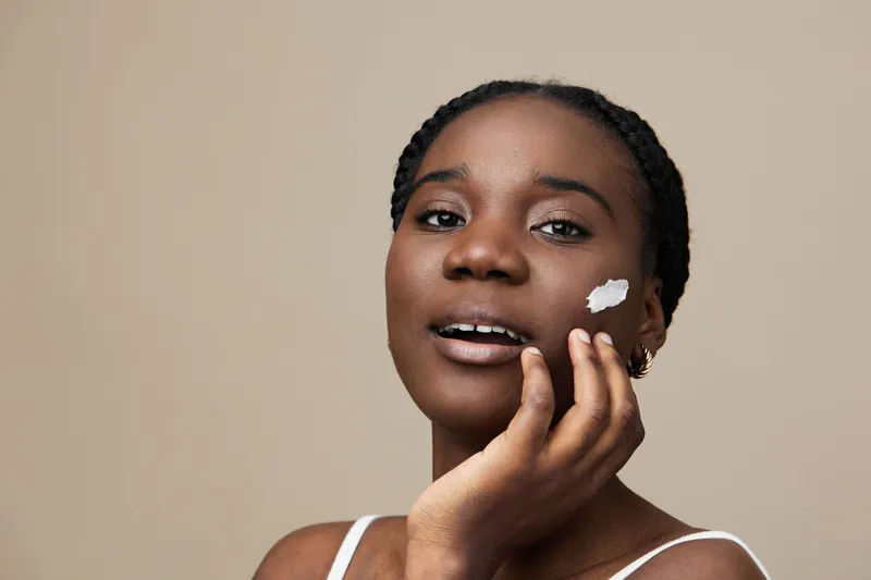Young African woman applying skincare cream on her face, showcasing healthy skin and beauty, with a warm neutral background enhancing the beauty concept