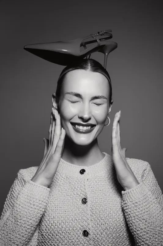 Happy caucasian woman with fair skin and closed eyes balances high heel shoe on head in studio.