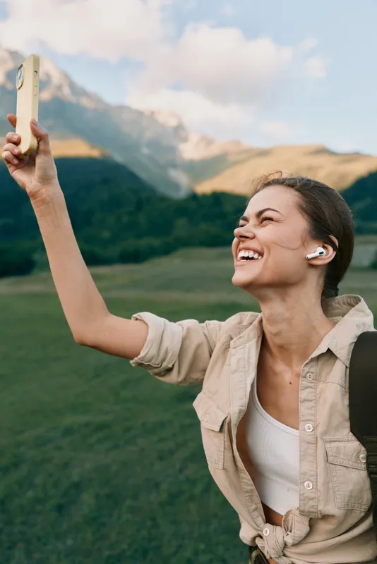 Happy woman outdoors taking selfie with smartphone, smiling, wearing wireless earbuds, casual outfit, mountain landscape background, natural light, lifestyle moment