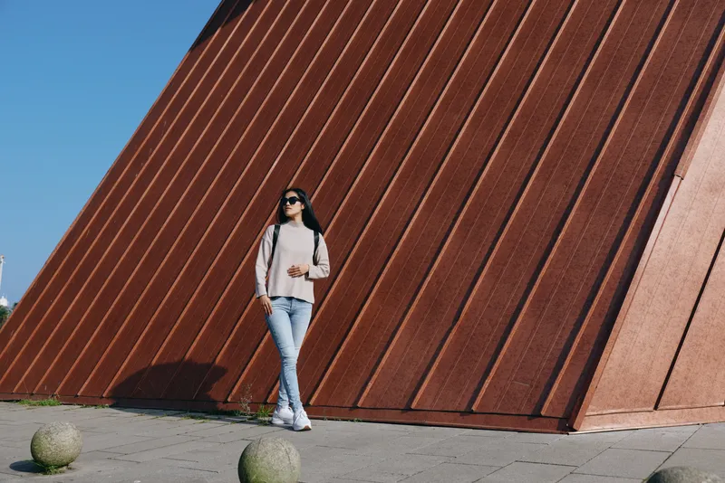 Outdoor fashion photo of a woman in jeans and casual top standing beside a large angular wall, sunglasses on, sunlight highlighting urban street style and confident pose