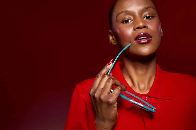 Confident African woman with dark skin wearing red sweater holds blue glasses near face on red background. Concept of fashion, beauty, style, and elegance with vibrant colors in studio.