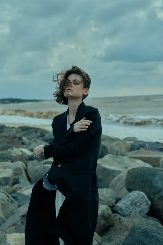 Woman in a dark coat stands on a rocky coast by the sea, wind tousling hair, moody editorial style for fashion, outdoor confidence and coastal drama.