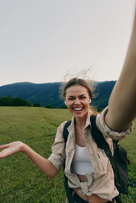 One person young woman with backpack takes a sunny meadow selfie with mountains behind, smiling broadly as a gentle breeze tousles hair, capturing adventure and carefree confidence