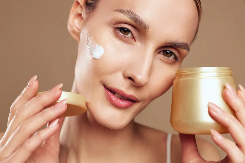 Young woman applies moisturizing cream on face holding open jar with cosmetic product skincare routine isolated on beige background closeup beauty treatment concept.