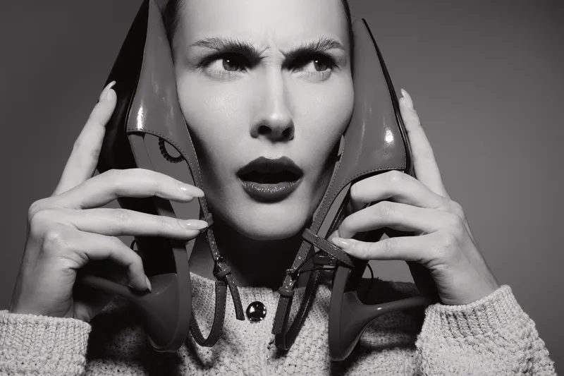 Confused white woman with light skin holds two red high heels near her ears, expressing surprise and doubt in black and white studio portrait.