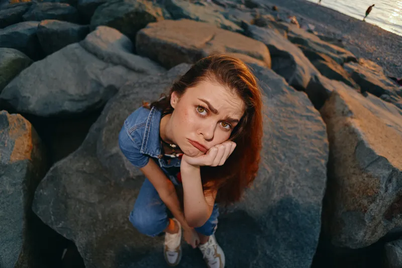Lifestyle woman in modern boho-western denim street style sitting thoughtfully on rocks by the sea with warm film color tones and casual outdoor vibe.