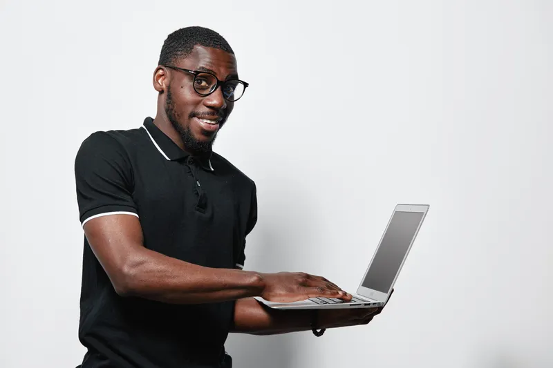 African American man with dark skin and glasses wearing black polo shirt holding laptop and smiling standing against white background for technology and communication.