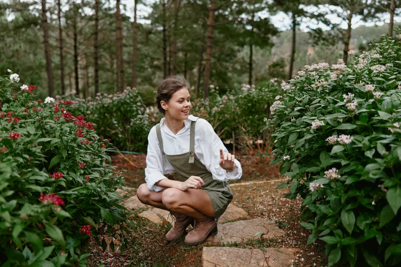 Young female gardener smiling among blooming flowers in a lush garden, wearing a white shirt and green overalls, showcasing a vibrant and seasonal atmosphere