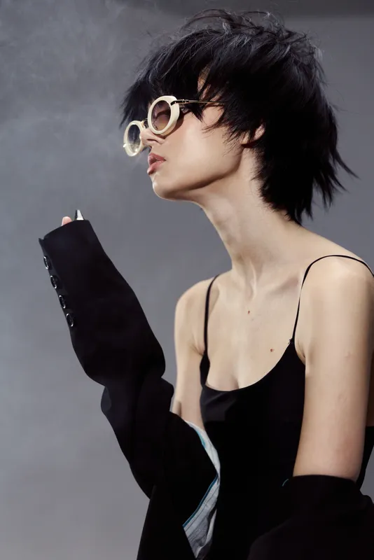 Stylish woman wearing oversized round sunglasses with short black hair and dark clothing against neutral background
