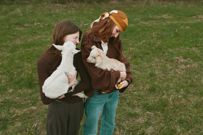 Two young women holding baby goats in a grassy field enjoy a relaxed lifestyle moment with warm clothing and natural smiles on a cloudy day outdoors.
